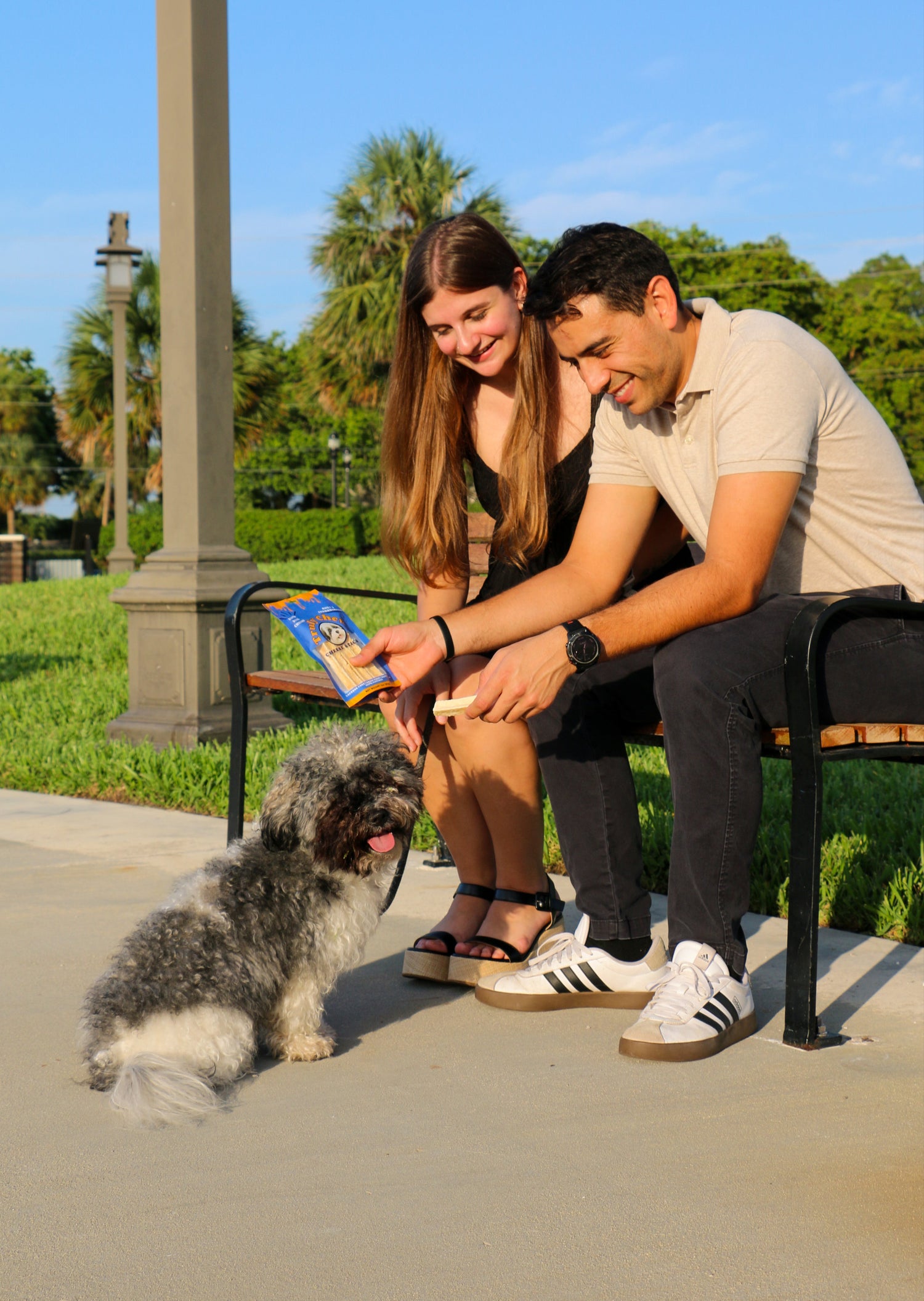 Couple sitting on a bench with a small dog, holding a bag of treats in a park setting.